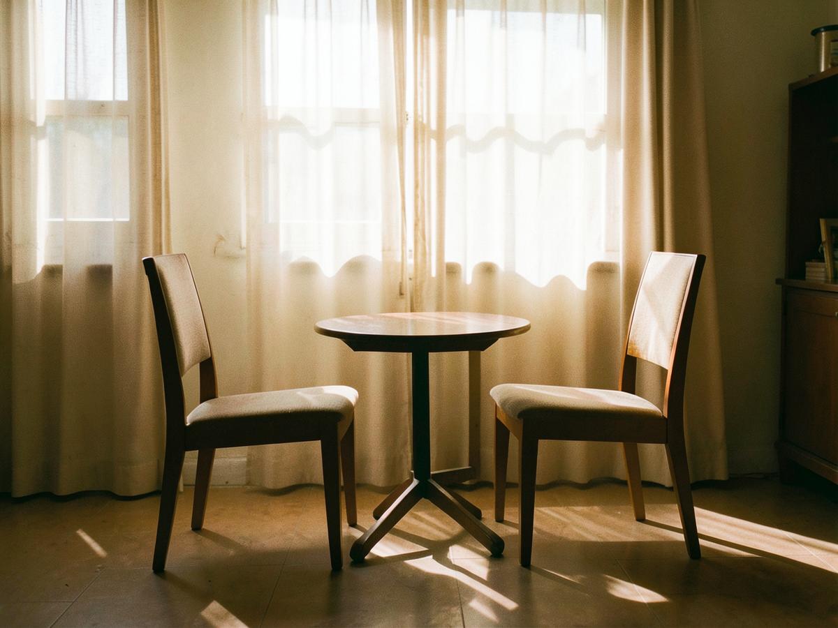 A small round table and two chairs beside a curtained window in afternoon light