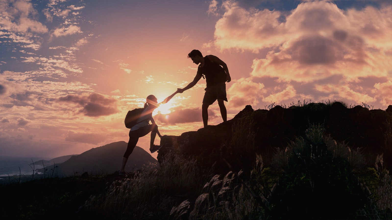 One person reaching down to help another up a ridge at sunrise